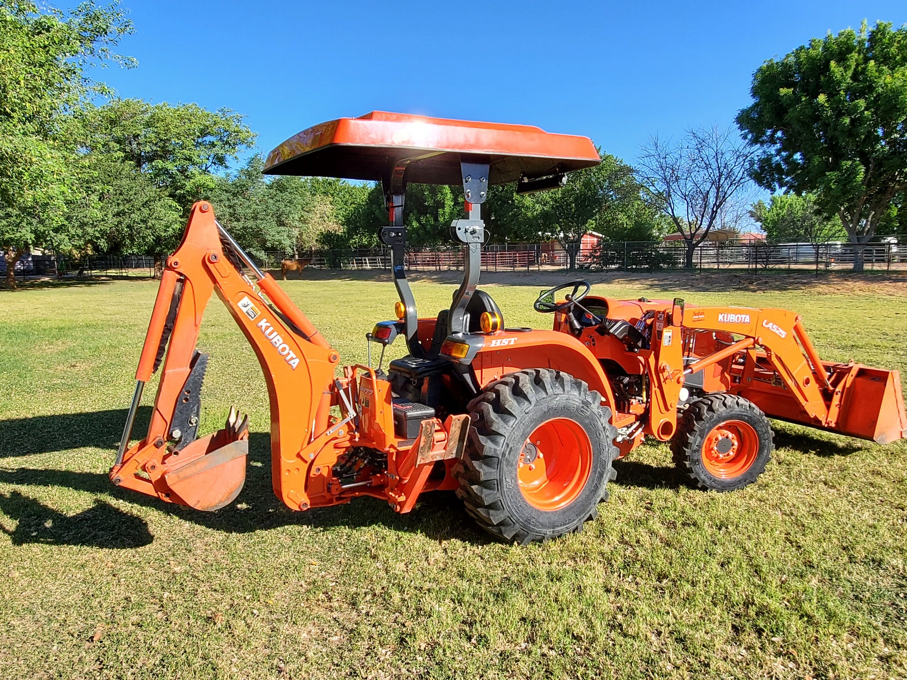 Rear three-quarter view of a Kubota compact tractor equipped with a front loader, backhoe, and orange Tractor Sunshade R-Series canopy.